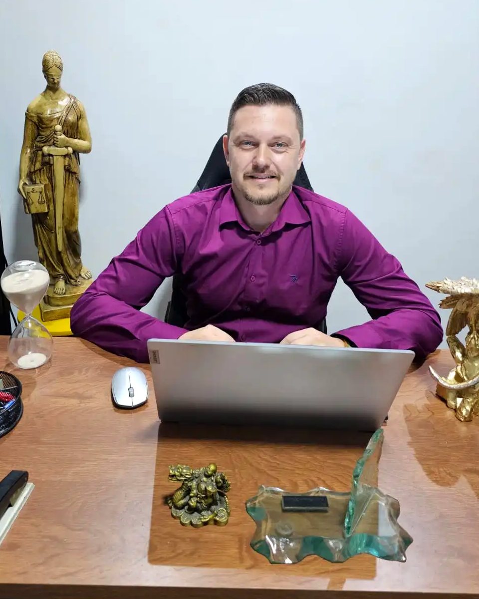 Dr. Marcos Eduardo Viveiro sentado à mesa do escritório, com notebook, estátua da Justiça e ampulheta