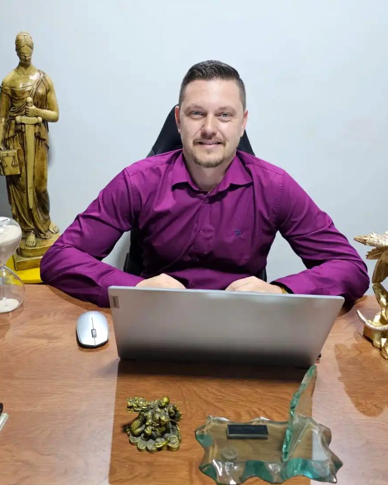 Dr. Marcos Eduardo Viveiro à mesa do escritório, sorrindo, com estátua da Justiça ao fundo